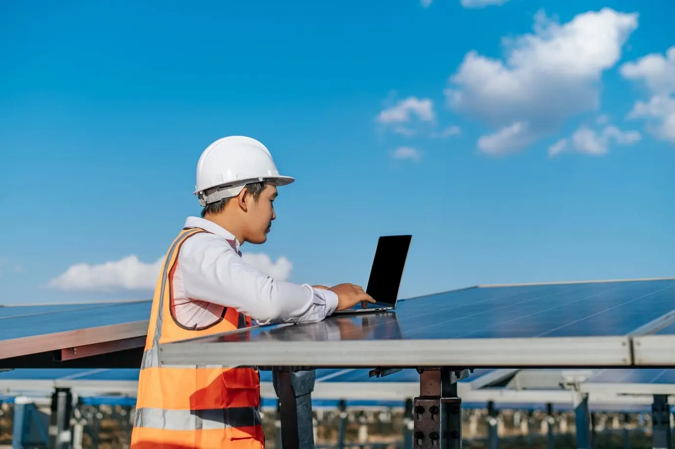 A young Asian inspection engineer and technician supervisor, male, wearing a white helmet, uses a laptop while working at a solar farm, checking sun and photovoltaic solar panel operation at the station, with copy space.